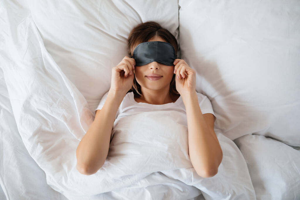 Agréable jeune femme couchée au lit et portant un masque oculaire avant d'aller dormir - Photo, image