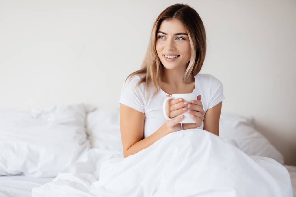 Radosna młoda dama spogląda w bok, relaksując się w łóżku z filiżanką herbaty - Zdjęcie, obraz