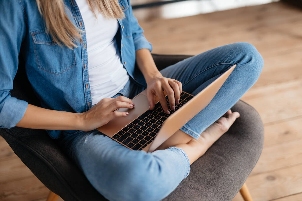 Gehakte foto van onherkenbare jongedame zittend met gekruiste benen in fauteuil en werkend aan haar laptop - Foto, afbeelding