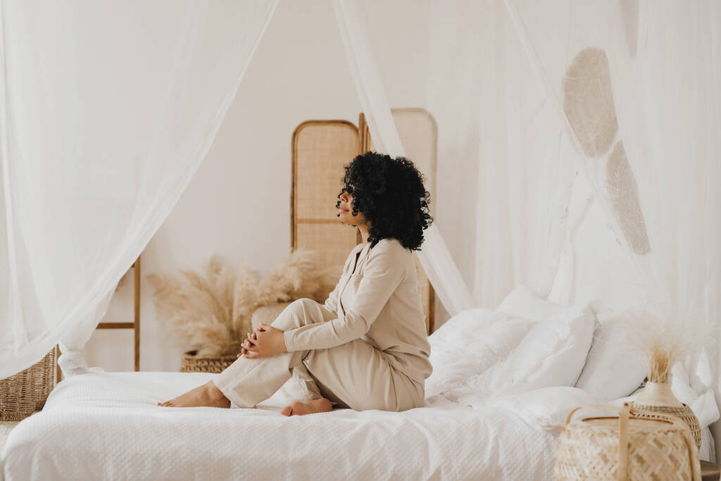 Krásná africká americká mladá žena sedí na posteli doma v bílém pyžamu. - Fotografie, Obrázek