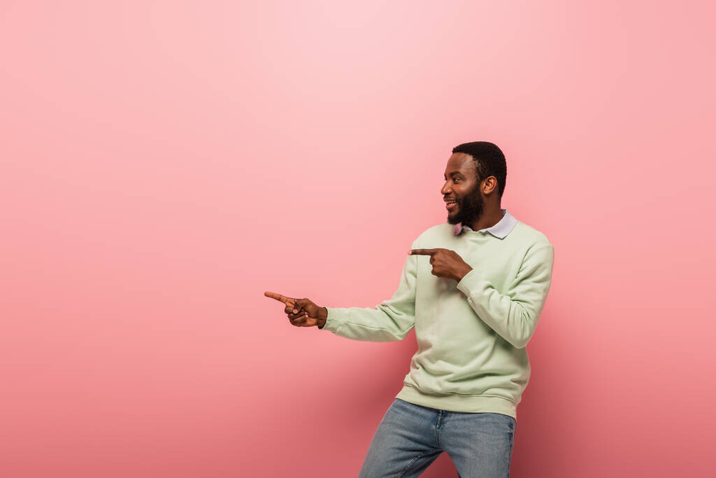 Vista lateral del hombre afroamericano sonriendo y señalando el fondo rosa  - Foto, imagen