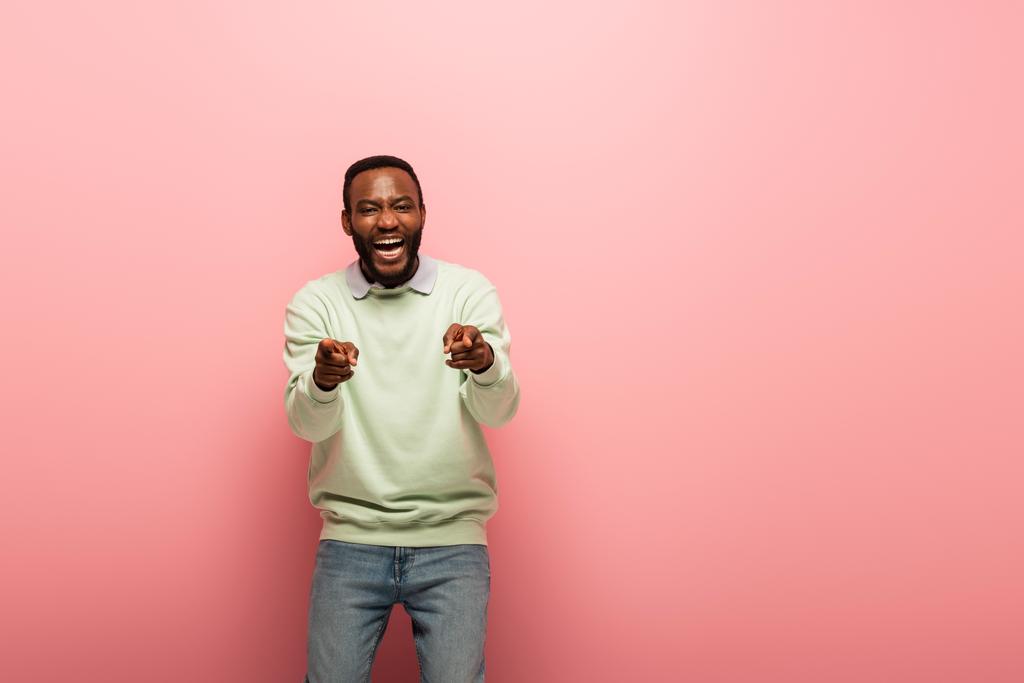 Веселый африканский американец, указывающий пальцами на камеру на розовом фоне  - Фото, изображение