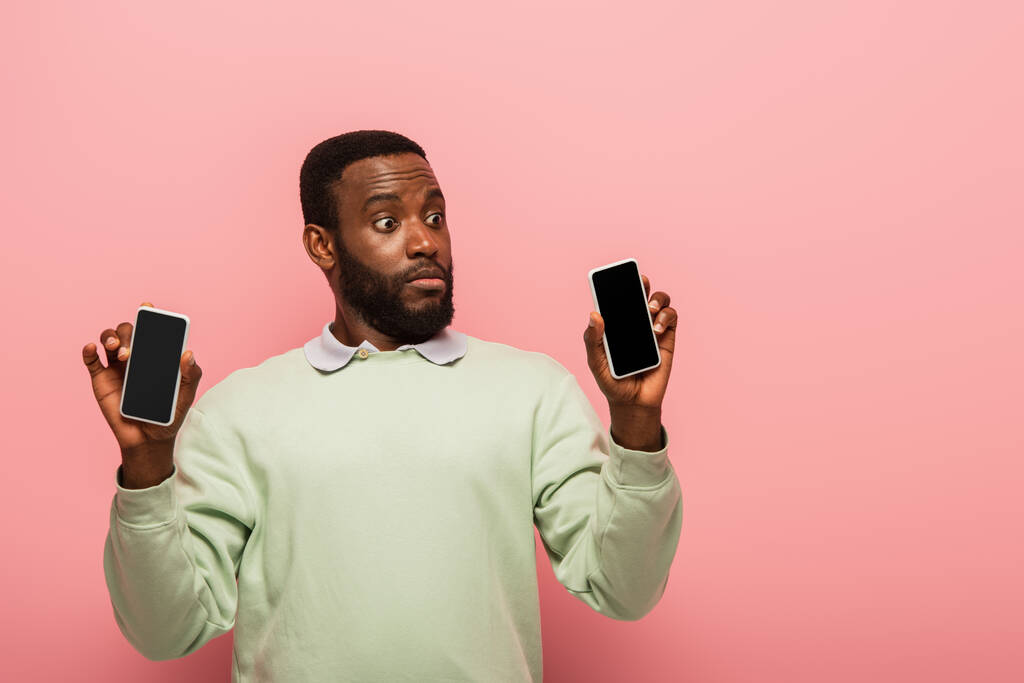 Zszokowany Afrykański Amerykanin trzyma smartfony na różowym tle  - Zdjęcie, obraz