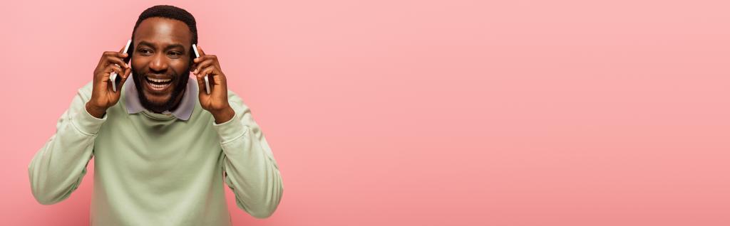 Hombre afroamericano positivo hablando en teléfonos celulares aislados en rosa, pancarta  - Foto, imagen