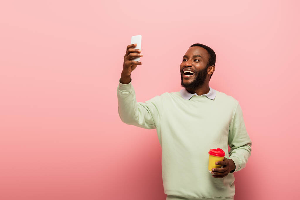 Sorrindo afro-americano homem com copo de papel tomando selfie no smartphone no fundo rosa  - Foto, Imagem