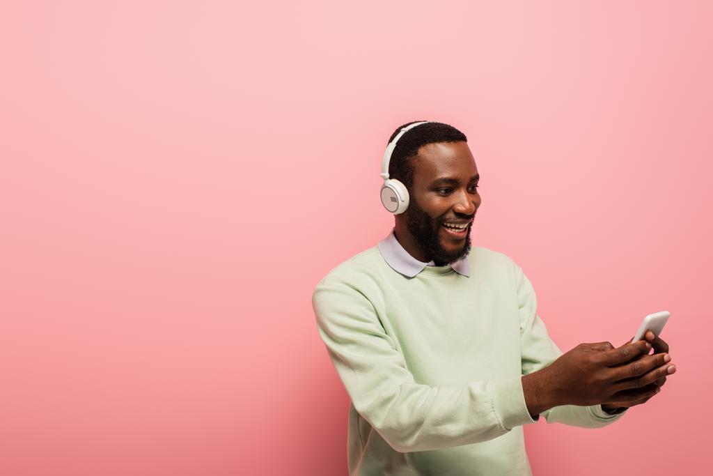 Usmívající se africký Američan ve sluchátkách pomocí smartphone na růžovém pozadí  - Fotografie, Obrázek