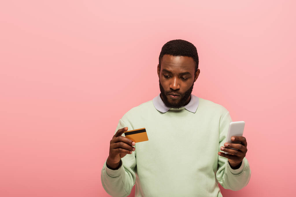 Afrikanischer Mann mit Kreditkarte und Smartphone beim Online-Shopping auf rosa Hintergrund  - Foto, Bild