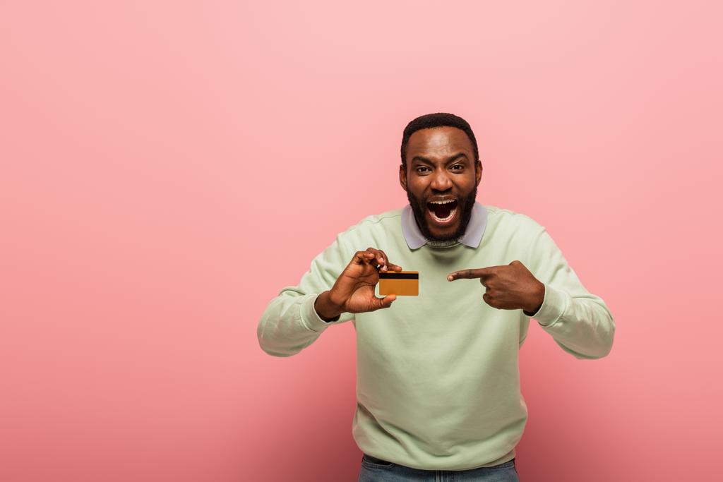 Σοκαρισμένος Αφροαμερικάνος που δείχνει με το δάχτυλο την πιστωτική του κάρτα σε ροζ φόντο  - Φωτογραφία, εικόνα