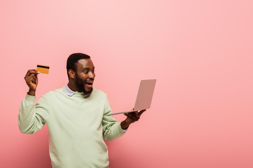 ピンクの背景にアフリカ系アメリカ人の男性がクレジットカードとノートパソコンを持っていて - 写真・画像