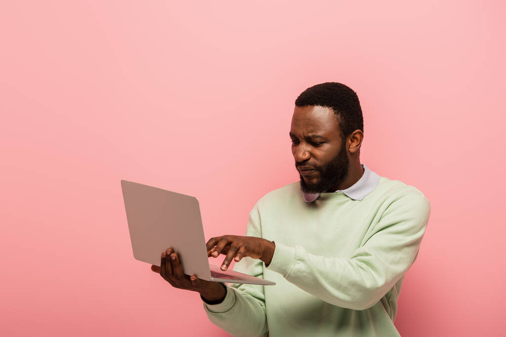незадоволений афроамериканський чоловік використовує ноутбук ізольований на рожевому
 - Фото, зображення