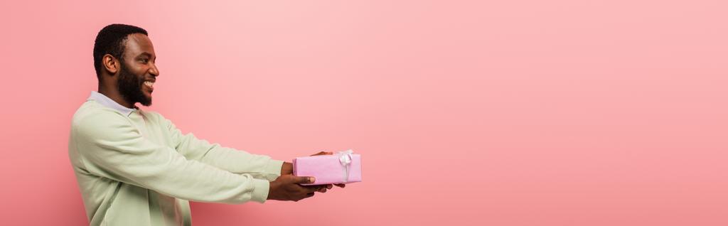 усміхнений афроамериканський чоловік тримає подарункову коробку в простягнутих руках ізольовано на рожевому, банер
 - Фото, зображення