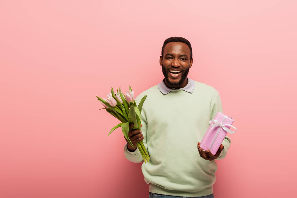 alegre afroamericano hombre con caja de regalo y tulipanes riendo de la cámara sobre fondo rosa - Foto, Imagen