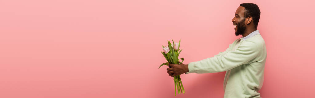 alegre hombre afroamericano sosteniendo tulipanes frescos en manos extendidas sobre fondo rosa, pancarta - Foto, imagen