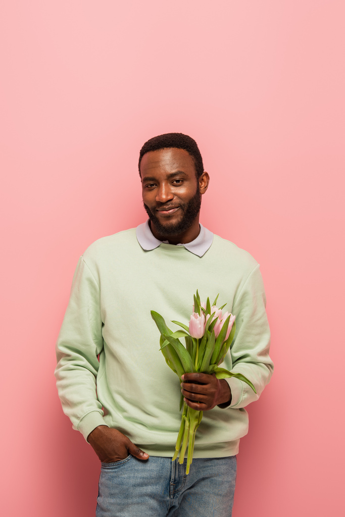 homme afro-américain avec des tulipes fraîches souriant à la caméra sur fond rose - Photo, image