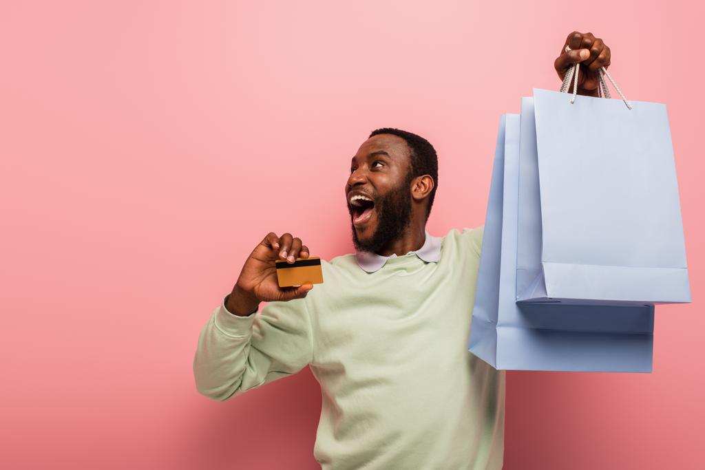 zdumiony Afrykański Amerykanin z kartą kredytową i torbami na zakupy patrząc na różowe tło - Zdjęcie, obraz