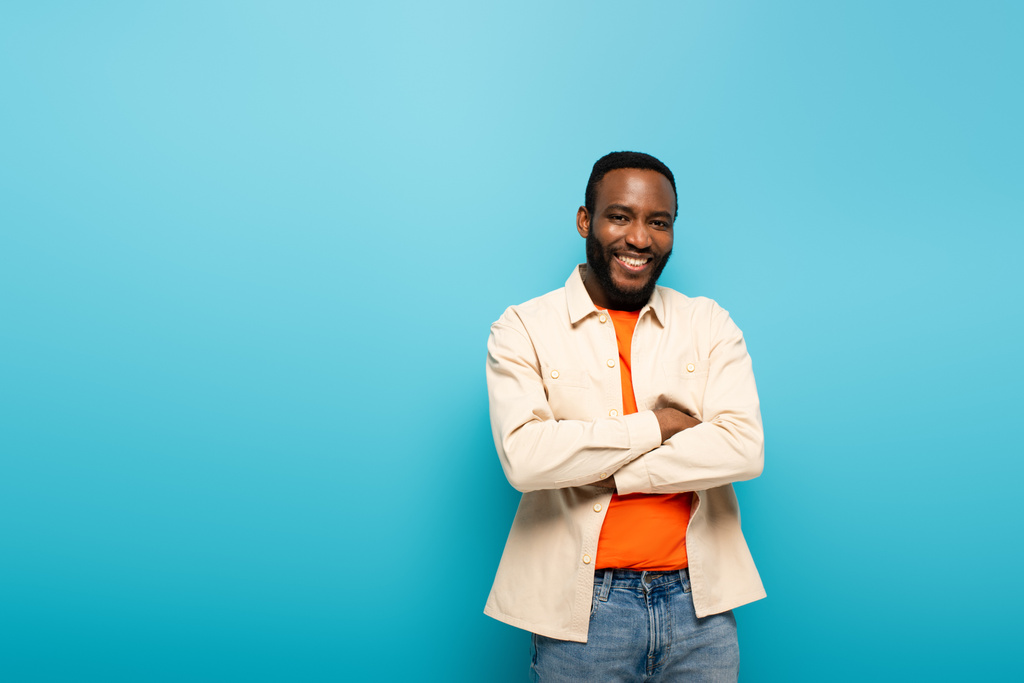 pozytywny Afroamerykanin uśmiecha się do kamery stojąc z skrzyżowanymi ramionami odizolowany na niebiesko - Zdjęcie, obraz