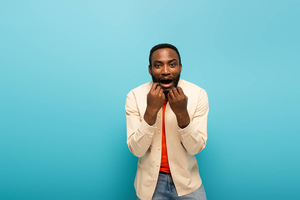 schockierter afrikanisch-amerikanischer Mann hält die Hände vor dem Gesicht, während er auf blauem Hintergrund in die Kamera schaut - Foto, Bild