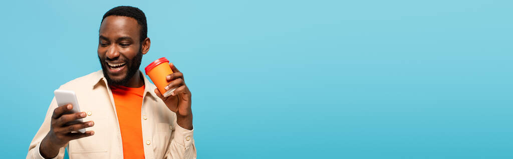 sonriente afroamericano hombre sosteniendo tarjeta de crédito mientras se utiliza el teléfono celular aislado en azul, bandera - Foto, Imagen