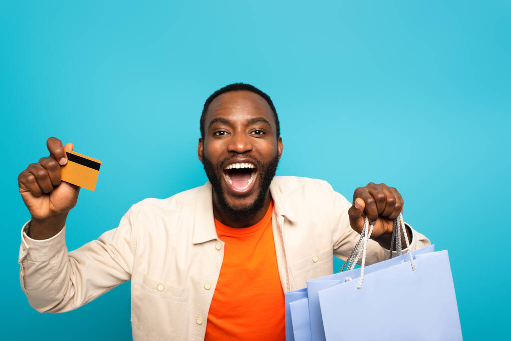 zdumiony Afroamerykanin człowiek z otwartymi ustami pokazując zakupy i karty kredytowej odizolowane na niebiesko - Zdjęcie, obraz