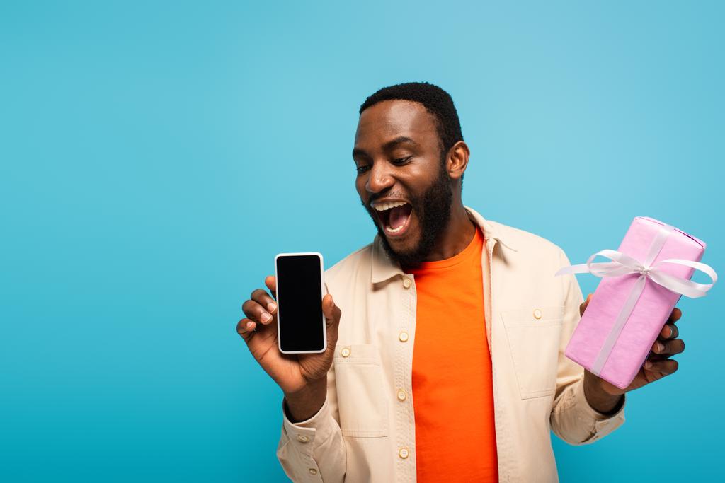 hombre afroamericano asombrado mostrando teléfono inteligente y caja de regalo aislado en azul - Foto, Imagen