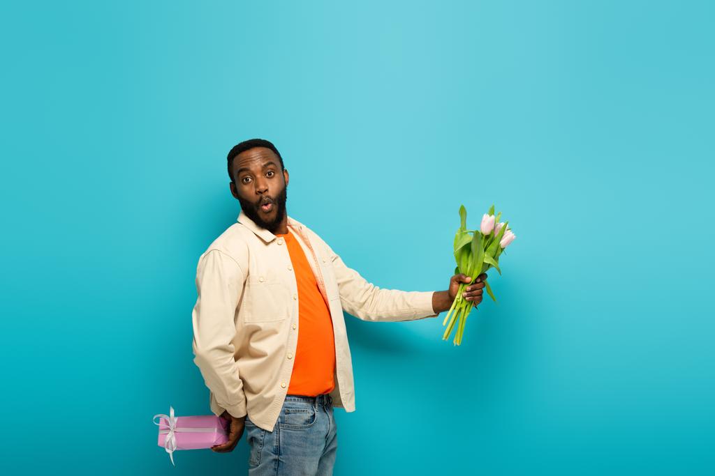 surpris homme afro-américain tenant tulipes fraîches et boîte cadeau sur fond bleu - Photo, image