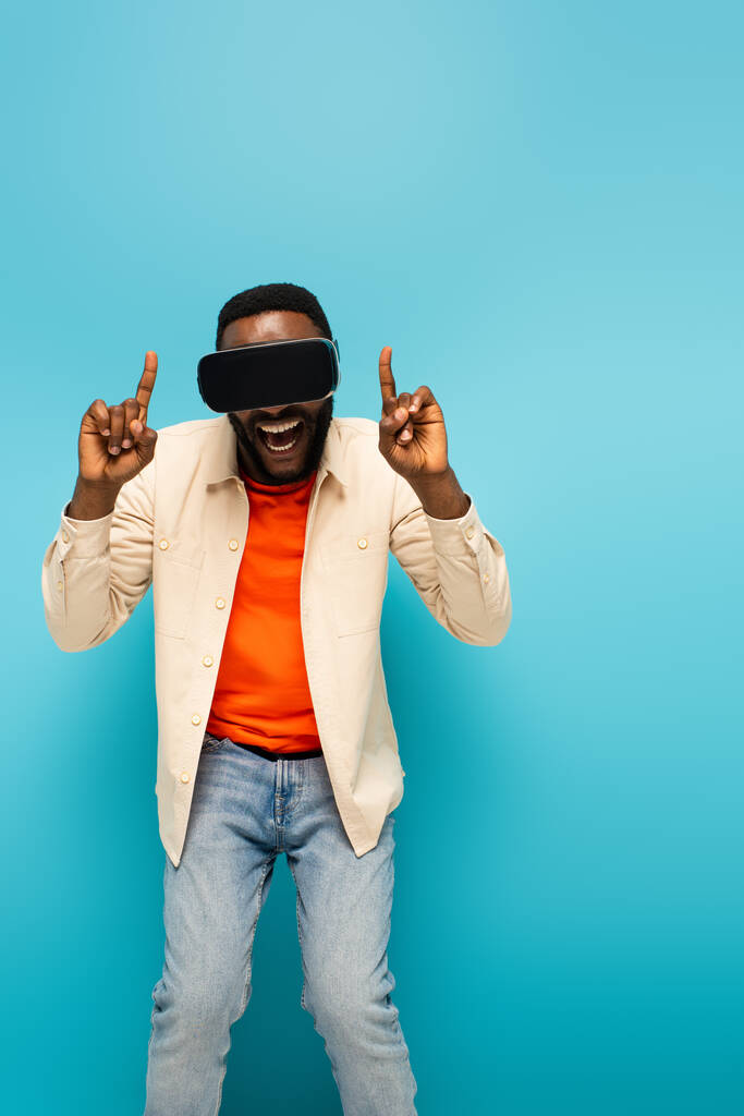 VR kulaklıklı afro-amerikan adam mavi arka planda parmaklarıyla işaret ediyor. - Fotoğraf, Görsel