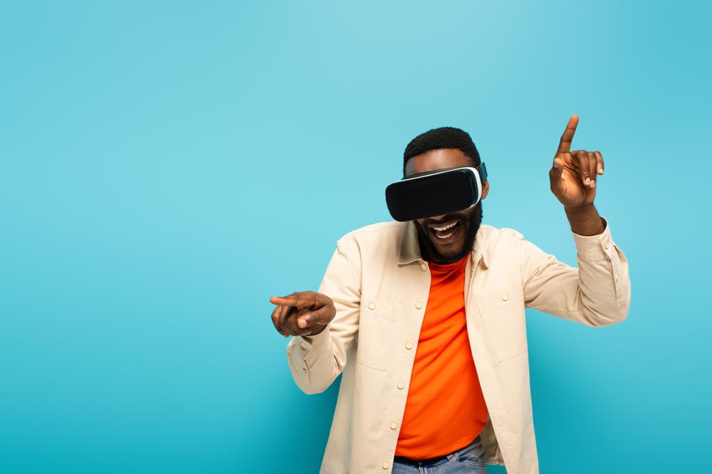fröhliche afrikanisch-amerikanische Mann in vr Headset zeigt mit Fingern isoliert auf blau - Foto, Bild