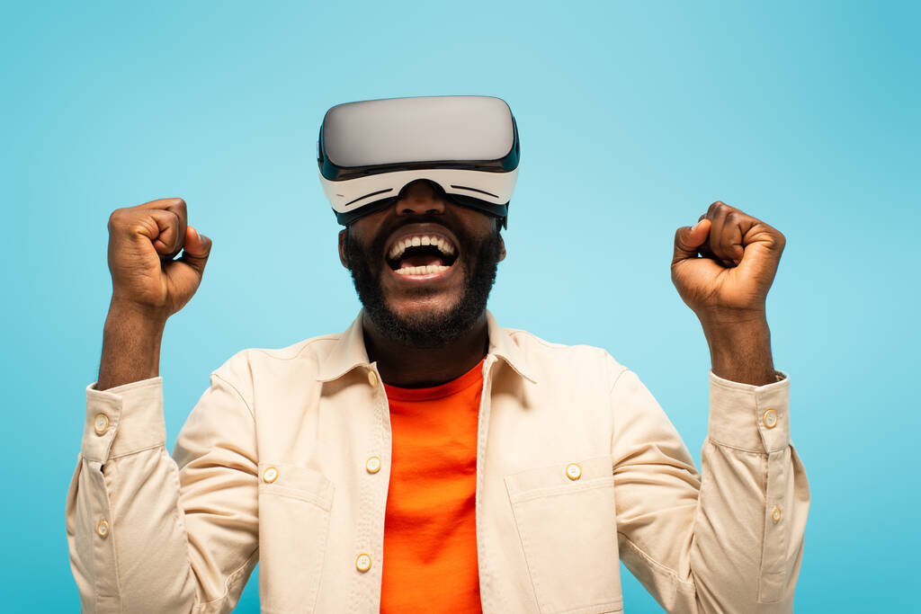 Африканский американец кричит и показывает жест успеха во время игры в VR гарнитуре изолированы на синий - Фото, изображение