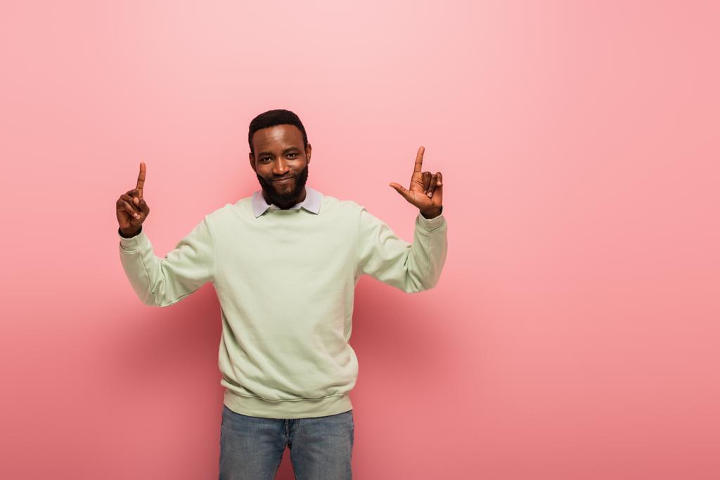 alegre afroamericano hombre sonriendo a la cámara y apuntando hacia arriba con los dedos sobre fondo rosa - Foto, Imagen