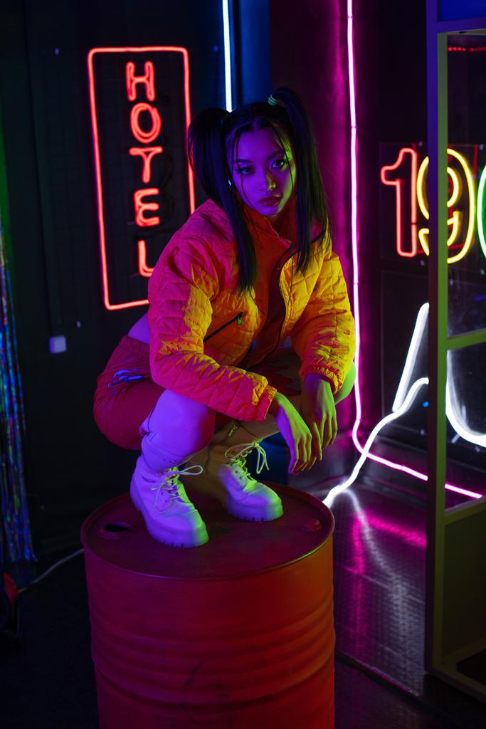 brunette asian woman sitting near neon sign with hotel lettering outside  - Zdjęcie, obraz