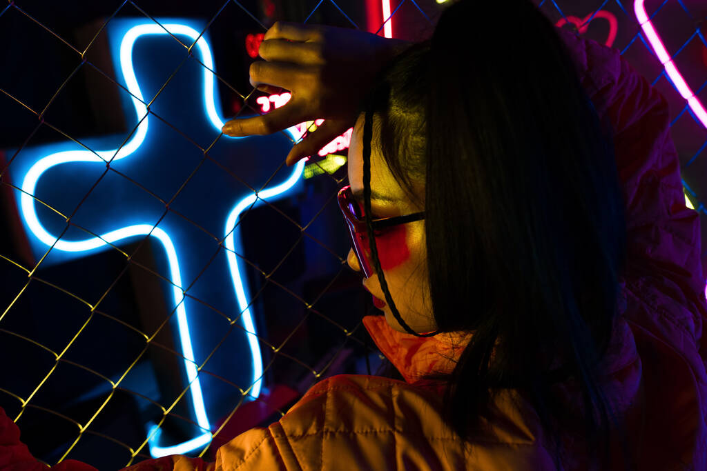стильная молодая азиатка в солнцезащитных очках, позирующая возле неонового знака и металлического забора  - Фото, изображение