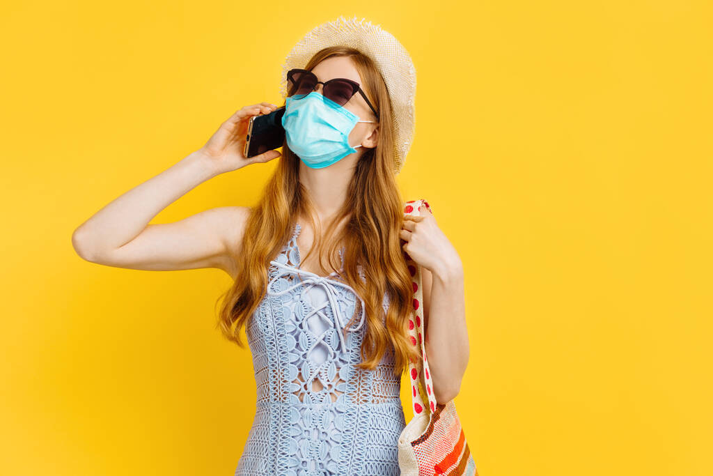 Молода щаслива жінка в медичній масці, в літній сукні і капелюсі розмовляючи на мобільному телефоні на жовтому тлі, концепцію літніх канікул - Фото, зображення
