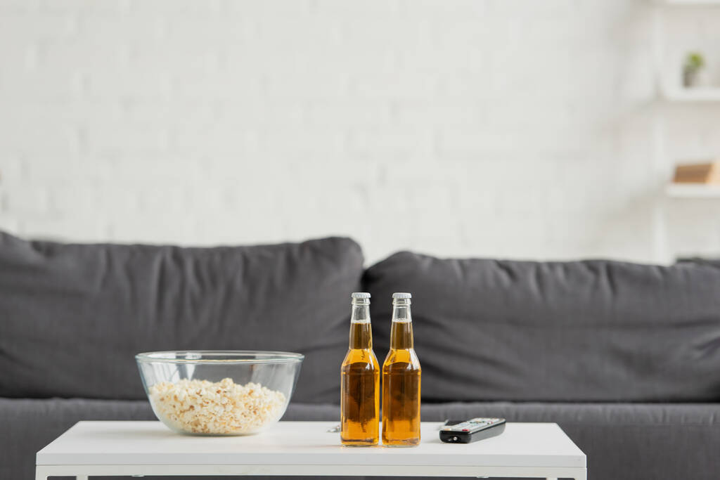 front view of two bottles of beer, popcorn and TV remote on blurred background with couch - Photo, Image