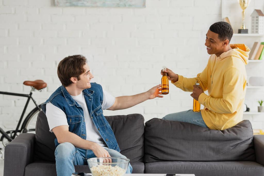 позитивные молодые межрасовые друзья пьют пиво вместе в гостиной - Фото, изображение