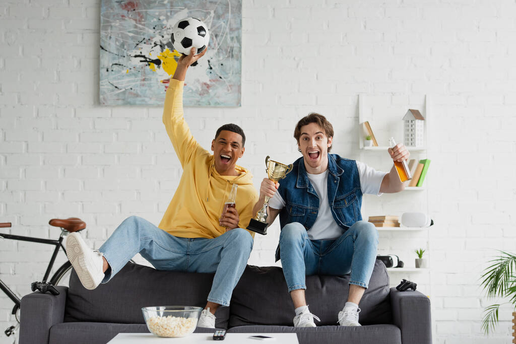 KYIV, UKRAINE - MARCH 22, 2021: cheerful interracial football fans sitting on couch and celebrating victory with beer and triumph cup in loft - Photo, Image