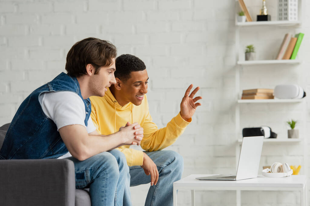 alegres amigos interracial sentados juntos y mirando en el ordenador portátil en la sala de estar moderna - Foto, imagen