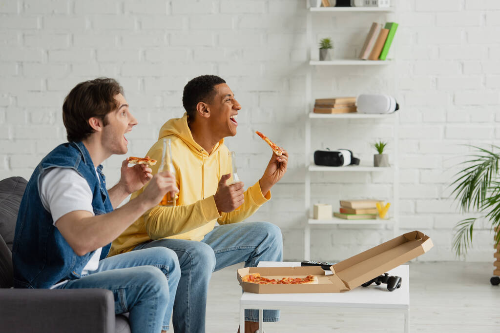 QUIIV, UCRÂNIA - MARÇO 22, 2021: amigos inter-raciais sentados no sofá com garrafas de cerveja e pizza e emocionalmente assistindo tv na sala de estar  - Foto, Imagem