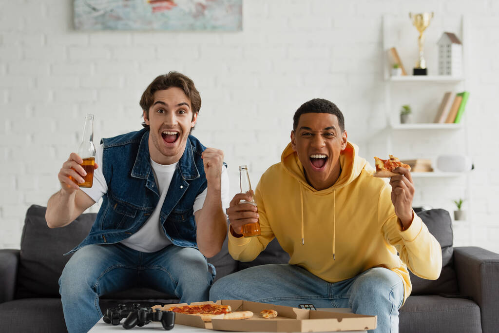 QUIIV, UCRÂNIA - MARÇO 22, 2021: amigos inter-raciais sentados no sofá com garrafas de cerveja e pizza  - Foto, Imagem