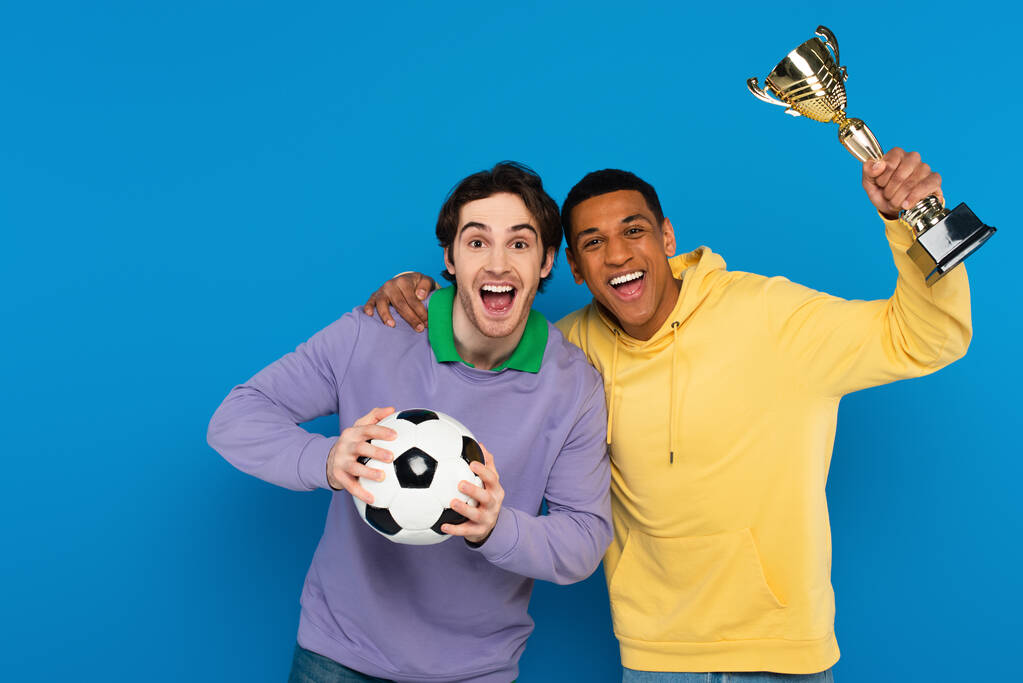 excited interracial football players holding ball and trophy cup isolated on blue - Photo, Image