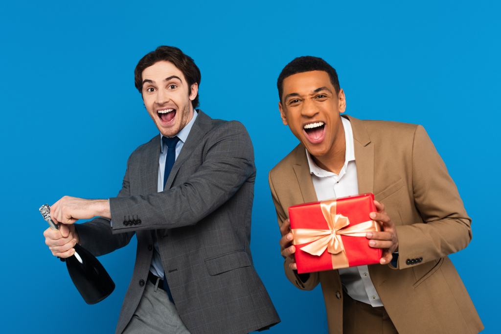 amigos interracial alegre en trajes divertirse con caja de regalo y botella de champán aislado en azul - Foto, Imagen