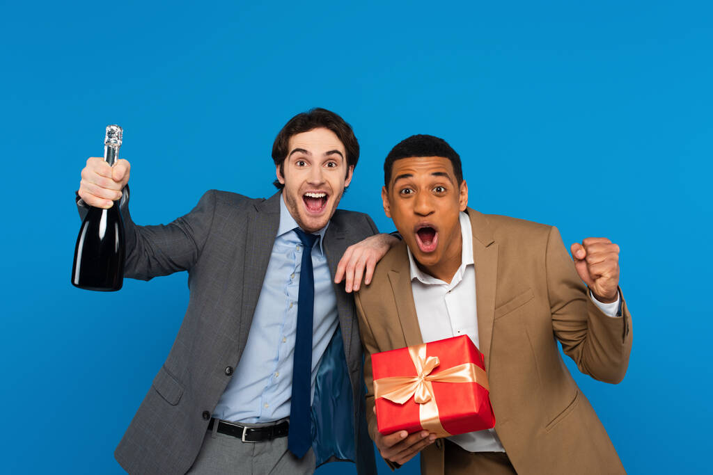 joyeux interracial amis en costumes souriant à la caméra et tenant boîte cadeau et bouteille de champagne isolé sur bleu - Photo, image