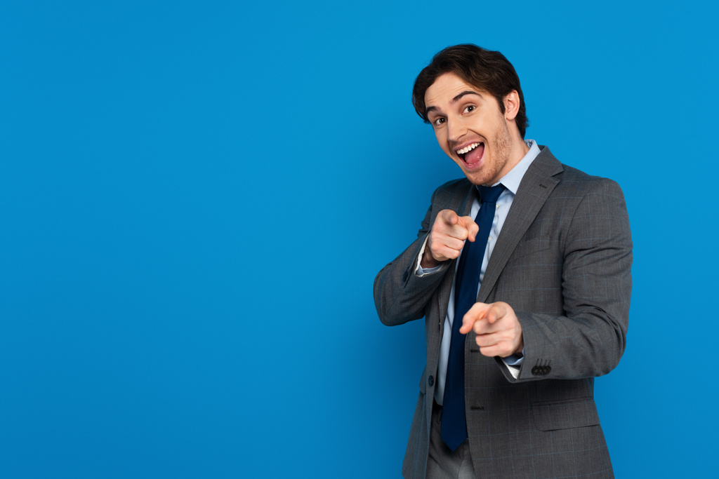 Fröhlicher junger Mann im eleganten Anzug zeigt mit den Fingern in die Kamera und lächelt vereinzelt auf blauem Grund - Foto, Bild