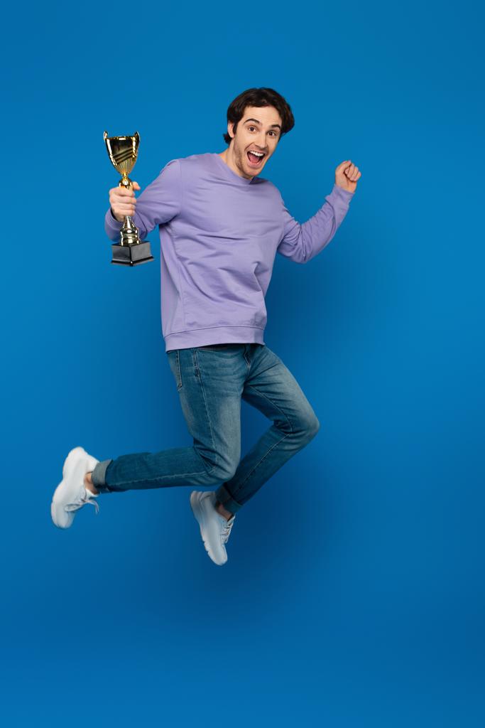glücklich lächelnder Mann in violettem Sweatshirt springt mit Pokal auf blauem Hintergrund - Foto, Bild