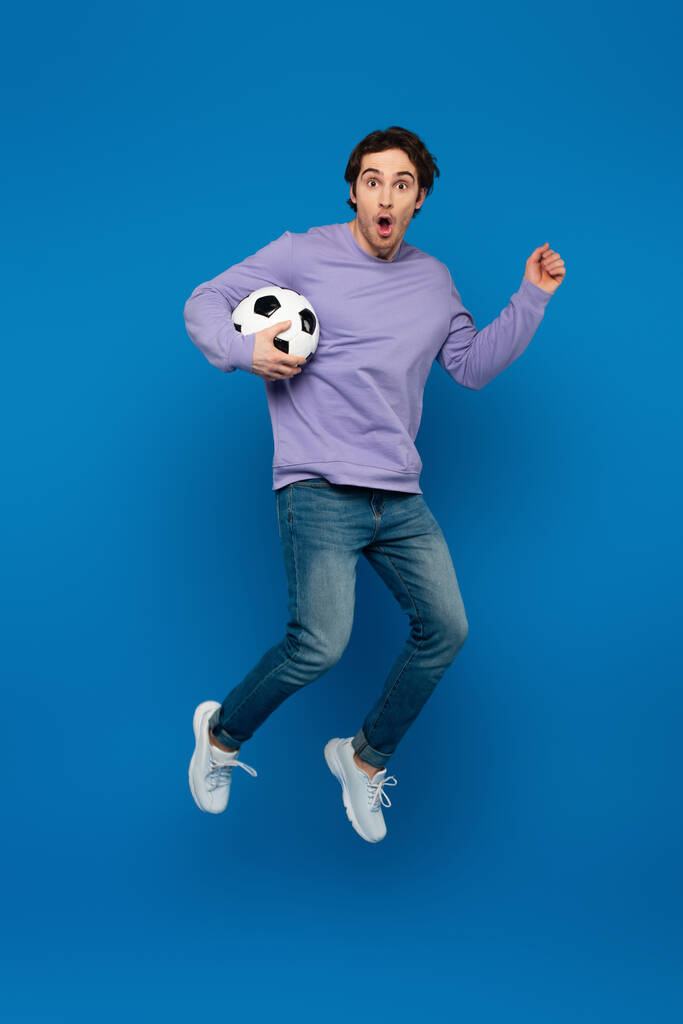 gelukkig lachende man in violette sweater springen met voetbal op blauwe achtergrond - Foto, afbeelding