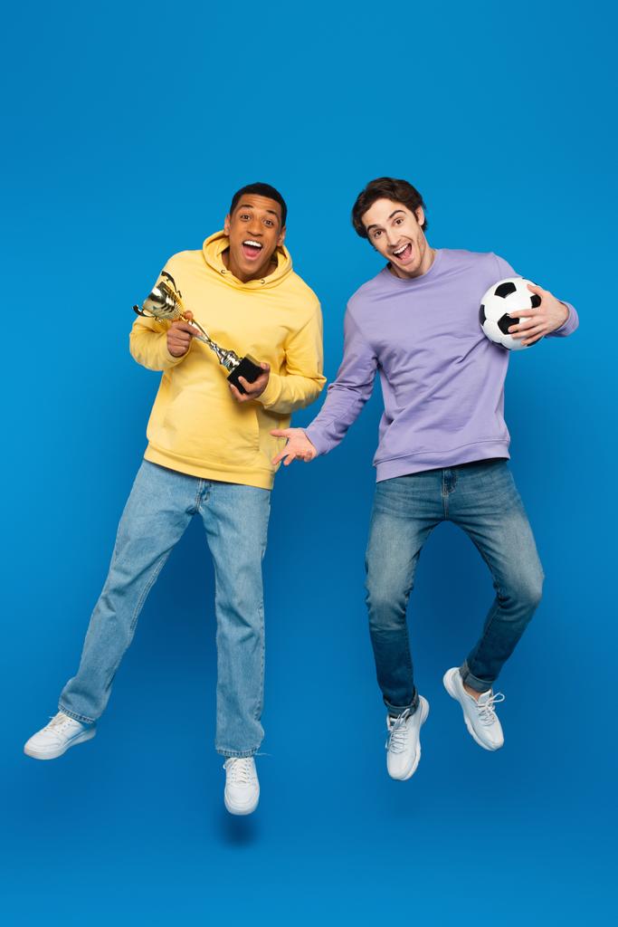 ευτυχισμένος Αφροαμερικάνος με φίλο αιωρείται και κρατώντας το ποδόσφαιρο και το κύπελλο στο μπλε φόντο - Φωτογραφία, εικόνα