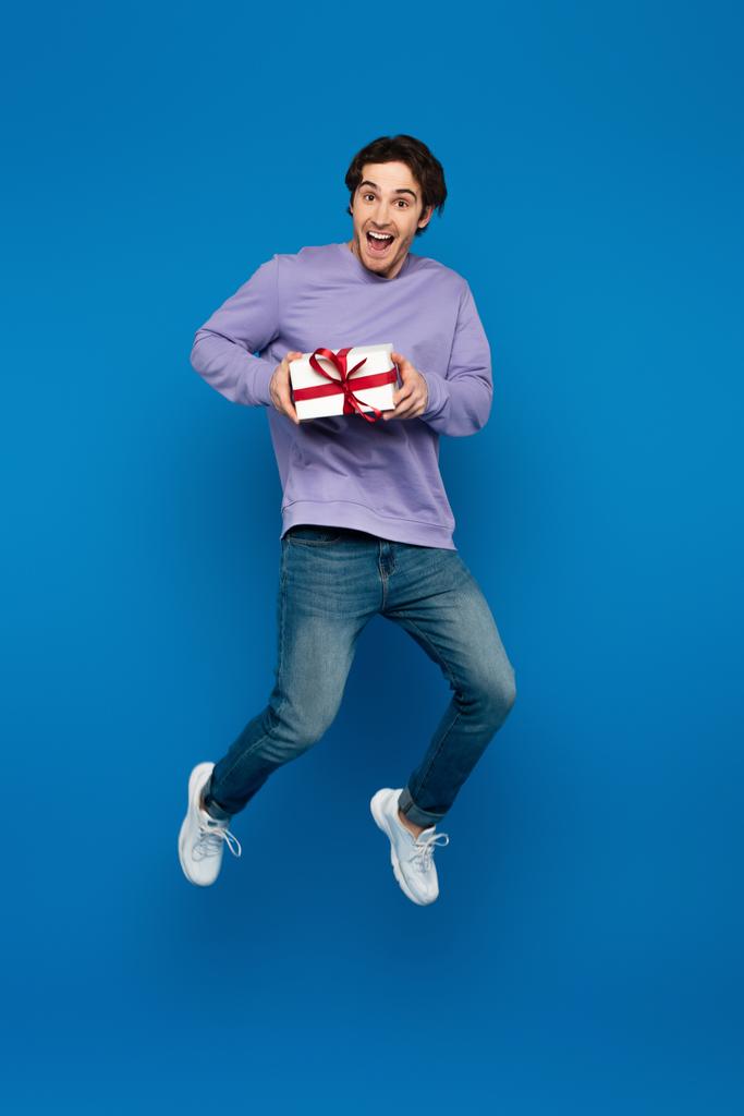 aufgeregter junger Mann schwebt mit Geschenkbox auf blauem Hintergrund - Foto, Bild