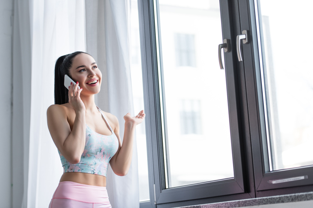 femme sportive souriant tout en parlant sur téléphone portable près de la fenêtre à la maison - Photo, image