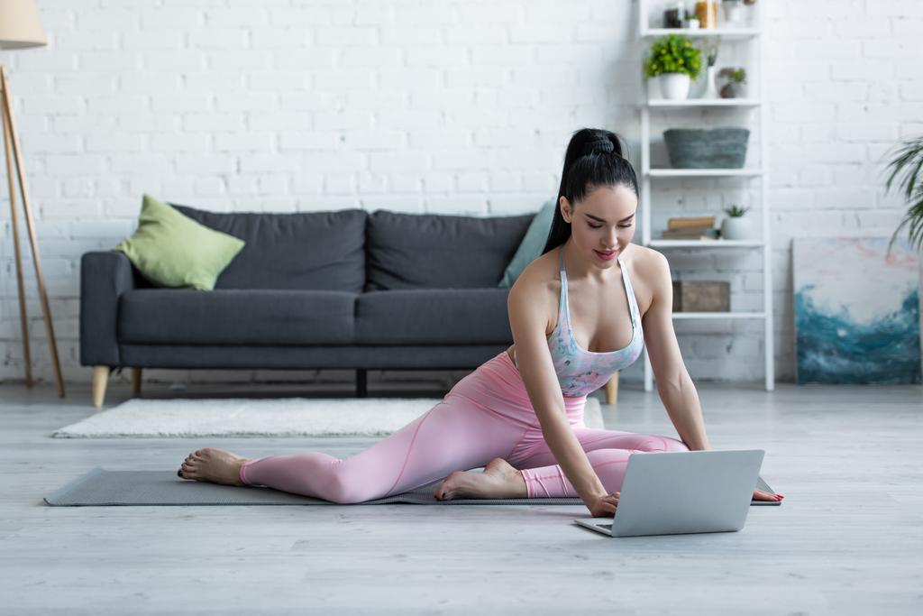 brünette Frau in Sportbekleidung schaut auf Laptop, während sie auf Yogamatte sitzt - Foto, Bild