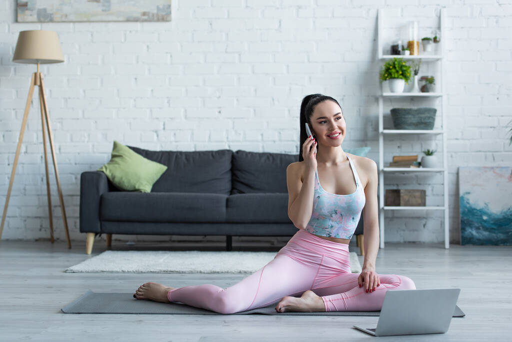 gelukkig jong vrouw praten op mobiele telefoon op yoga mat in de buurt laptop - Foto, afbeelding
