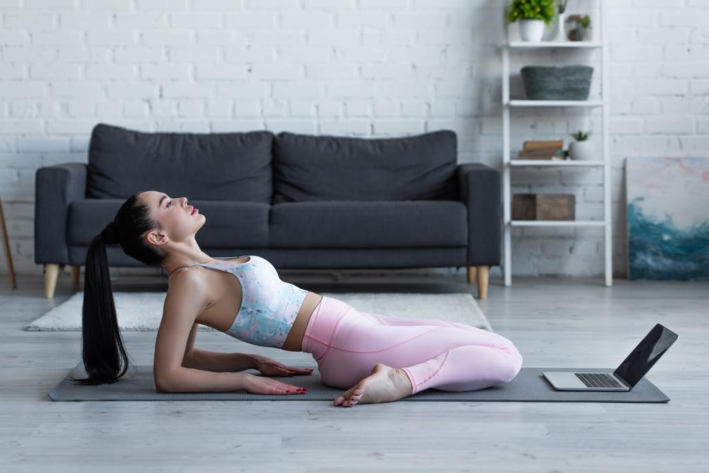 side view of woman practicing hero pose on yoga mat near laptop - Photo, Image